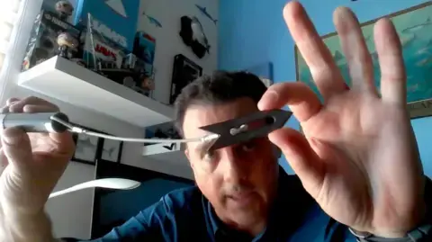 ماوریسو هوئوس برچسب صوتی را نشان میدهد؛ سیلندری است که به یک کابل متصل است و سر فلزی از آن آویزان میشود