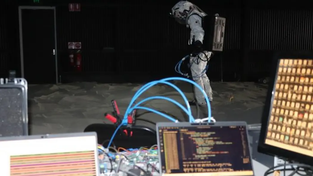 شخصی پوشیده در یک لباس سفید پشت میزی ایستاده که سیمها و رایانههای مختلفی روی آن قرار دارند