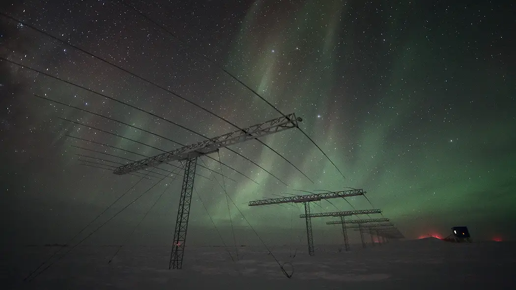 سلسله‌ای از سازه‌هایی که شبیه تیرهای برق به نظر می‌رسند، در سطح یخ‌زده به سمت افق امتداد دارند. آسمان بالای آن پر از ستارگان و با شفق‌های سبز خیره‌کننده پر شده است.