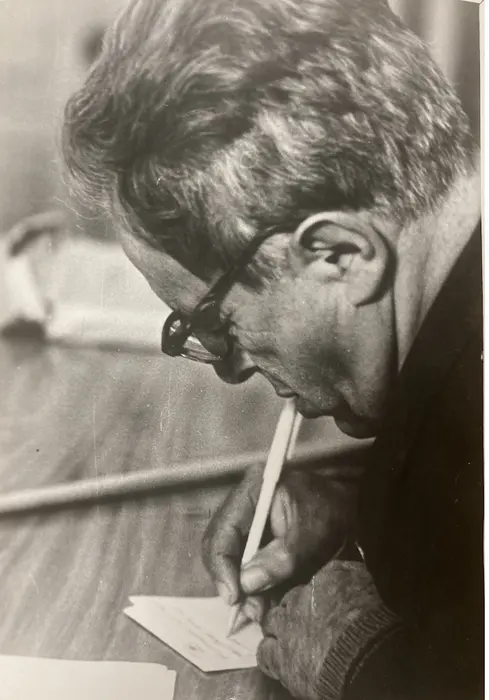 مردی پشت میز نشسته و با قلم در حال نوشتن است.