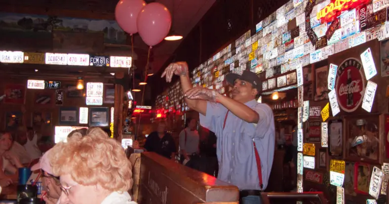 مردی با پیراهن دکمه‌دار راه‌راه آبی و سفید، بند شلوار قرمز و کلاه آبی در حال پرتاب یک نان در سالن غذاخوری کافه لمبرت است.