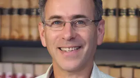 رولاند برون لبخند می‌زند و مستقیم به دوربین نگاه می‌کند. او موهای کوتاه خاکستری دارد و عینک با قاب سیمی بر چهره دارد. در پس‌زمینه کتاب‌هایی روی یک قفسه دیده می‌شود.