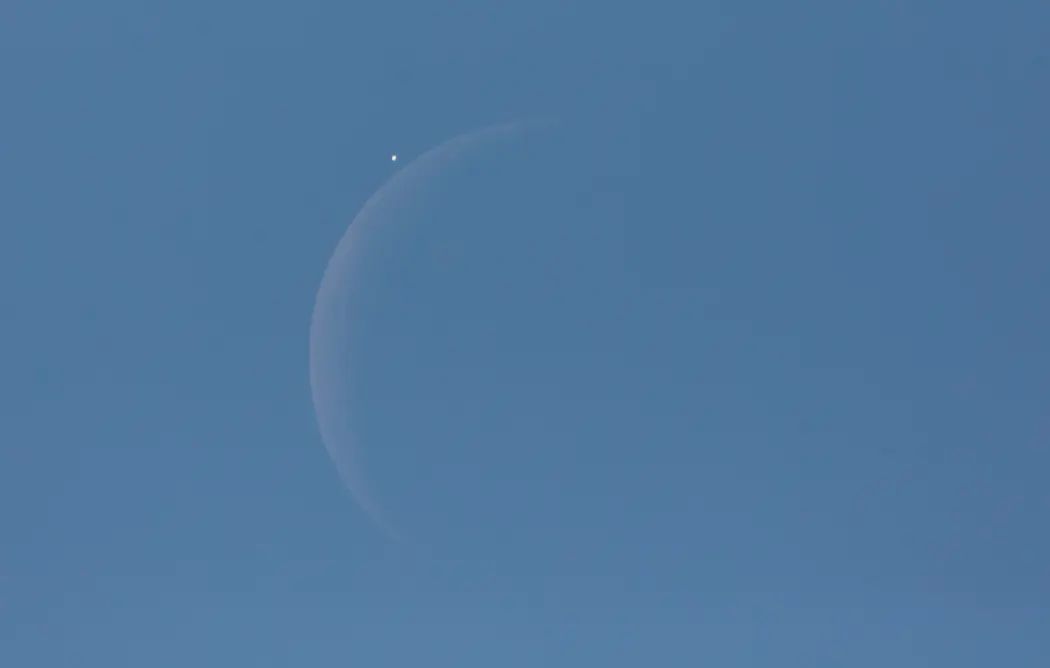 زهره در کنار ماه هلالی در طول روز، مقابل آسمان آبی، پیش از شروع مخفی شدن دیده میشود.