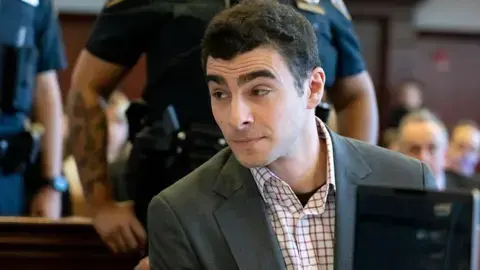 گتی ایمیجز؛ مانگیونه که بلیزر و پیراهن دکمه‌دار پوشیده، در دادگاه به طرفی نگاه می‌کند؛ پلیس پشت سر او ایستاده است