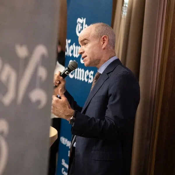 جو کان، در کت و شلوار آبی تیره، به میکروفون صحبت می‌کند. پشت سر او بنر آبی نشانگر لوگوی نیویورک تایمز را نمایش می‌دهد.