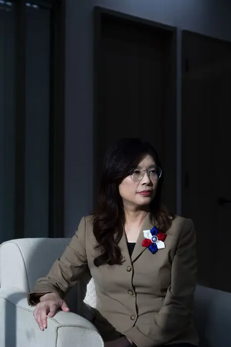 شخصی با موهای تیره و عینک، بر روی یک صندلی روشن نشسته است و ژاکت بژ به تن دارد؛ یک پین رزت با رنگ‌های سرخ، سفید و آبی بر یقه‌اش چسبیده است.