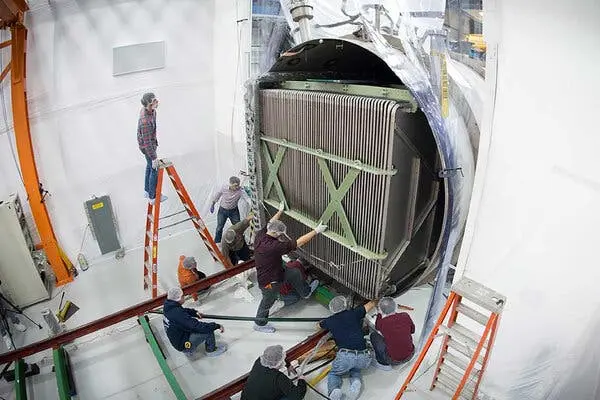 تصویری از بالا که چند کارگر با پوشش موهای سر، قطعه بزرگ مکعبی از تجهیزات علمی را داخل یک محفظه می‌گذارند.